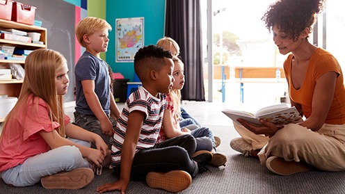 Students sitting in class while lady teacher reads from a book in a kindergarten classroom.