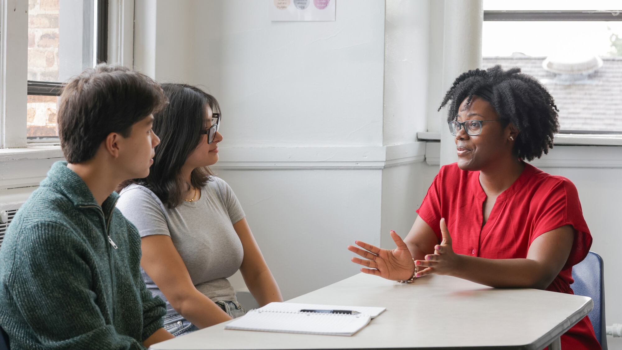 An African American female teacher talks with two teenage students at a table while wearing casual clothing.