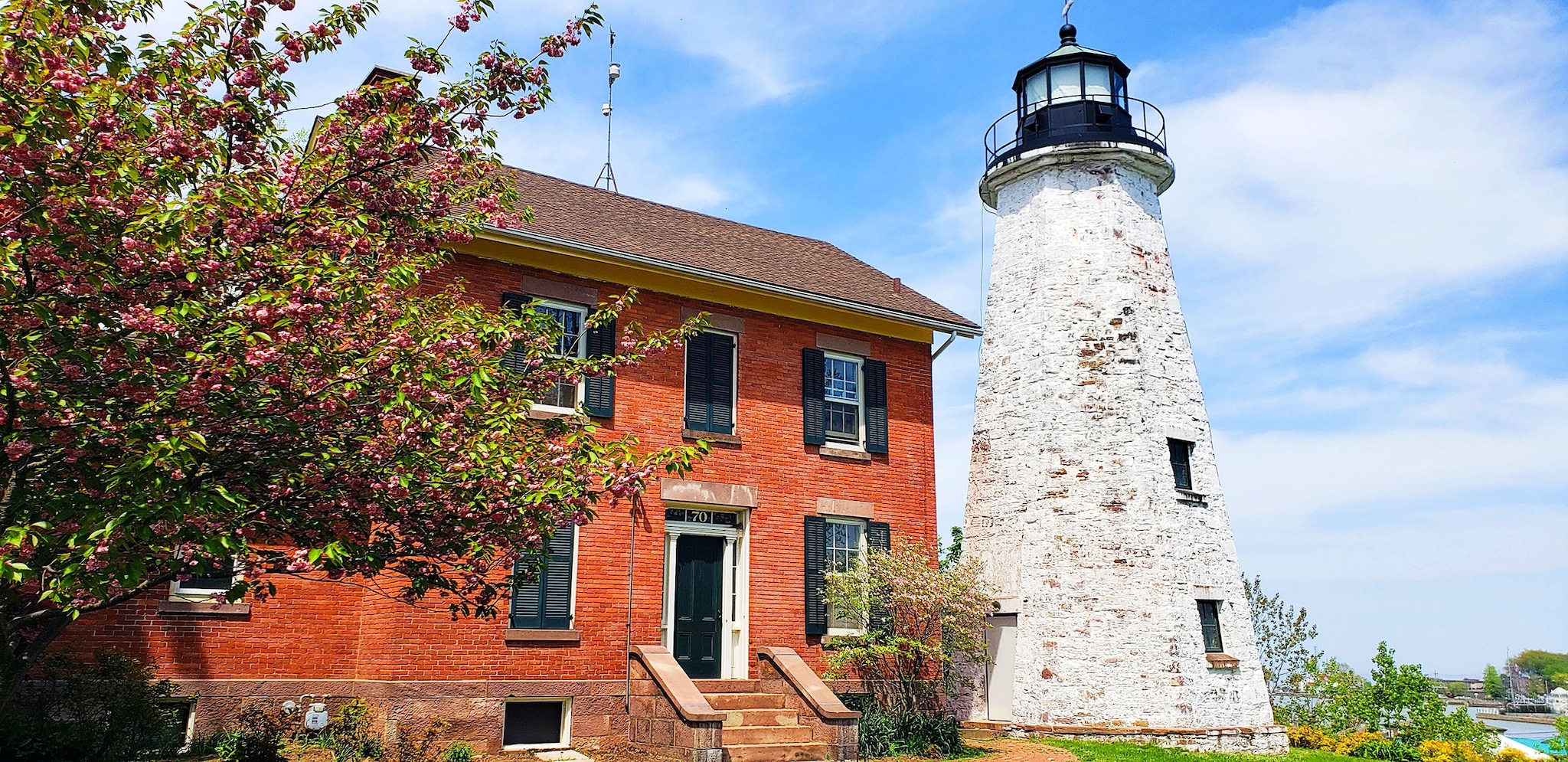 A red brick building with black shutters next to an old white lighthouse with a black top, surrounded by blooming trees and a clear blue sky.