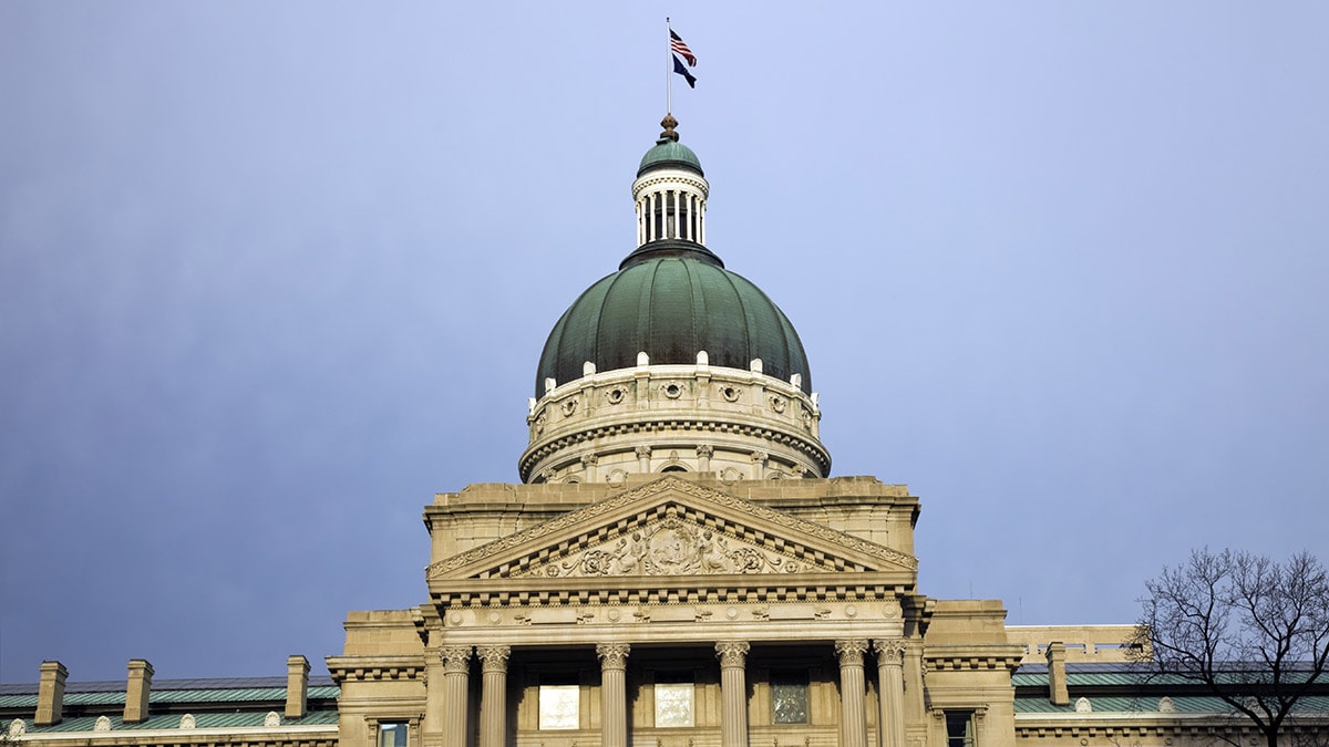 The Indiana state capitol building, featuring a large green dome and ornate stone facade under a cloudy sky.