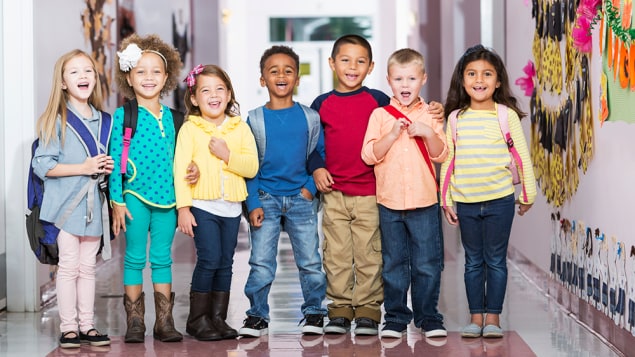 A group of seven children standing in a row in a school hallway, laughing and smiling at the camera. The little boys and girls are kindergarten or preschool age, 4 to 6 years.