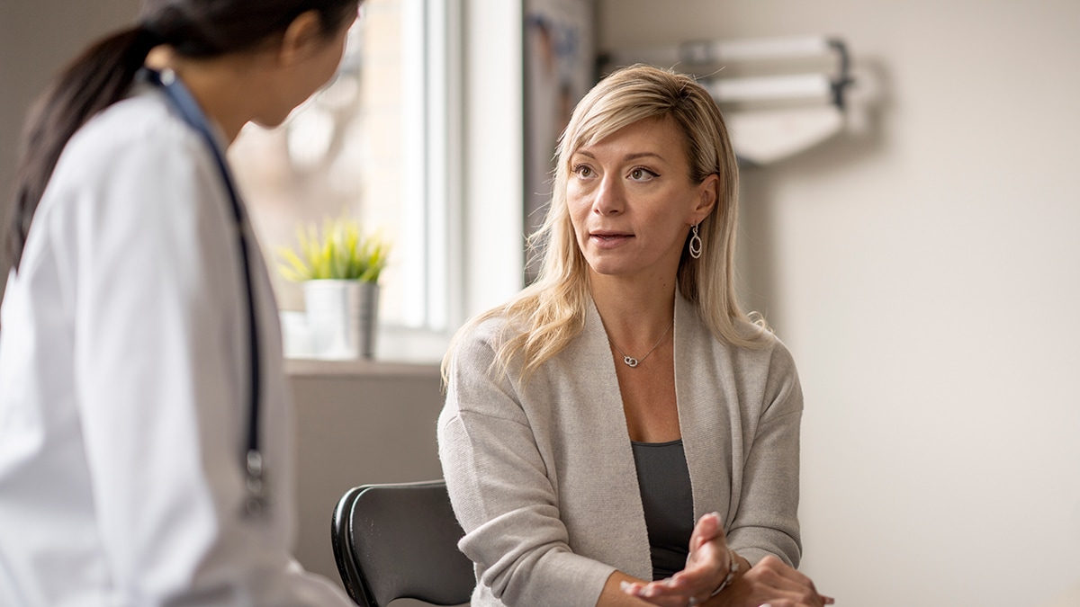 A middle aged woman sits down with her doctor to discuss her symptoms. She is dressed in casual business attire and looking at her doctor as they talk. The female doctor is wearing a white lab coat and listening attentively to the woman's concerns.