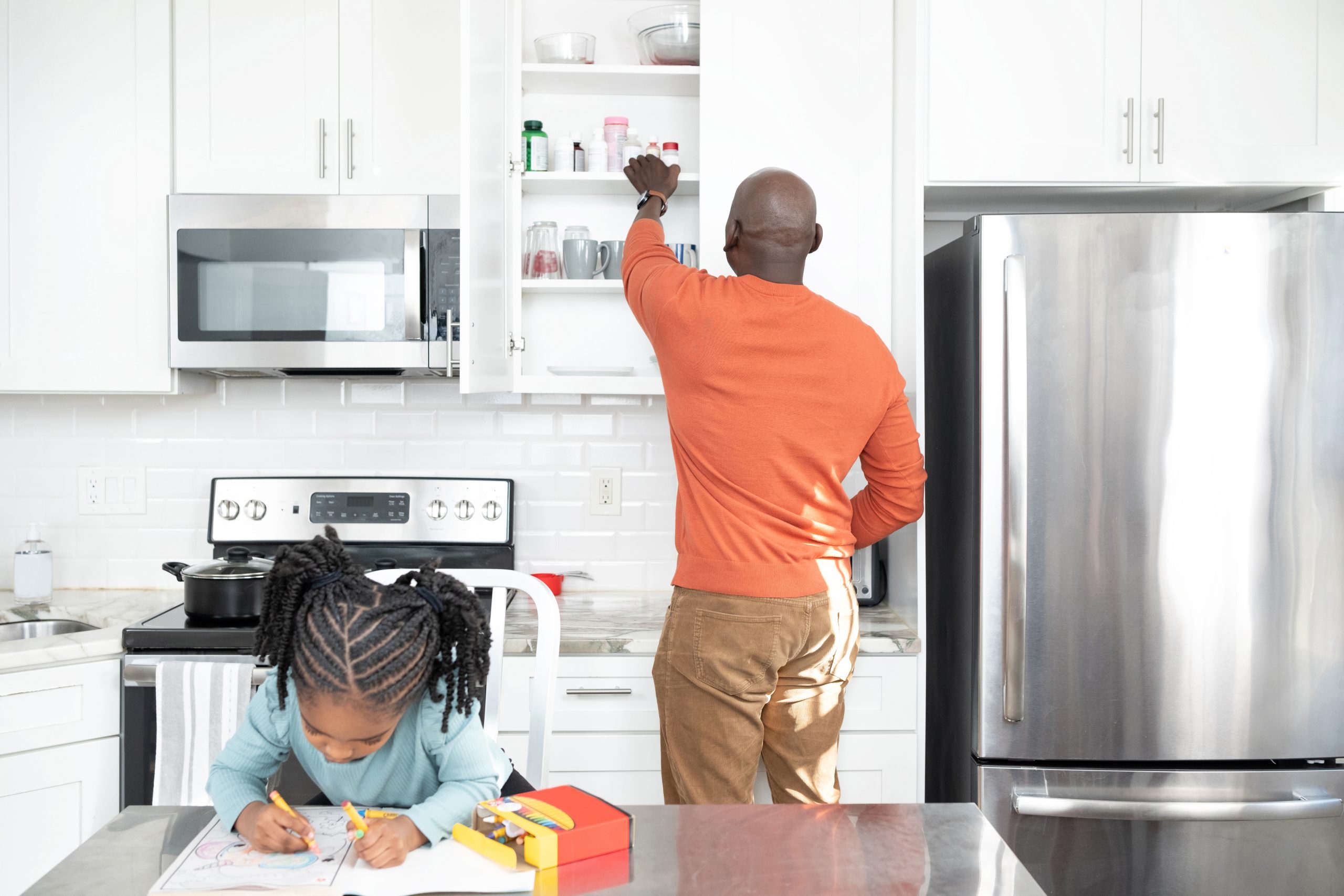 A kitchen scene with a young girl sitting at a table coloring and a father putting medicines in a high cabinet