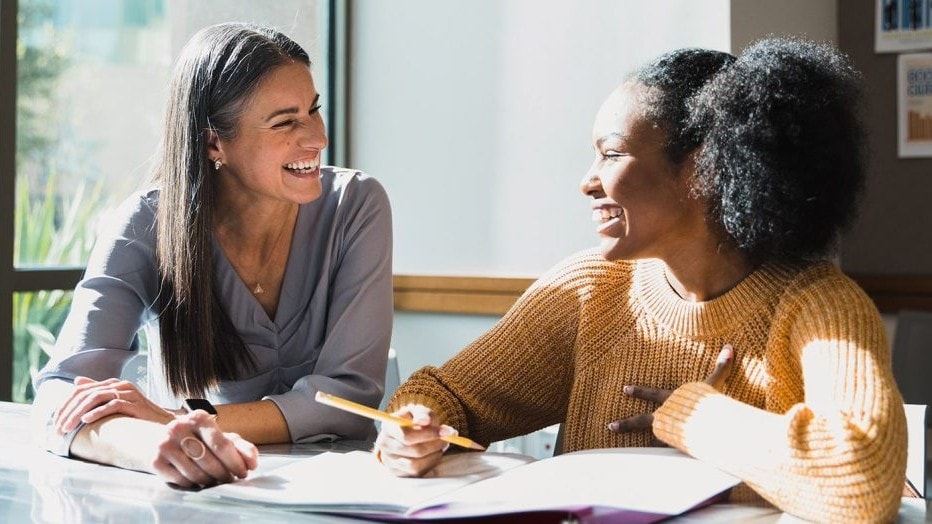 An adult helping a teenager with schoolwork, as they look at each other and smile.