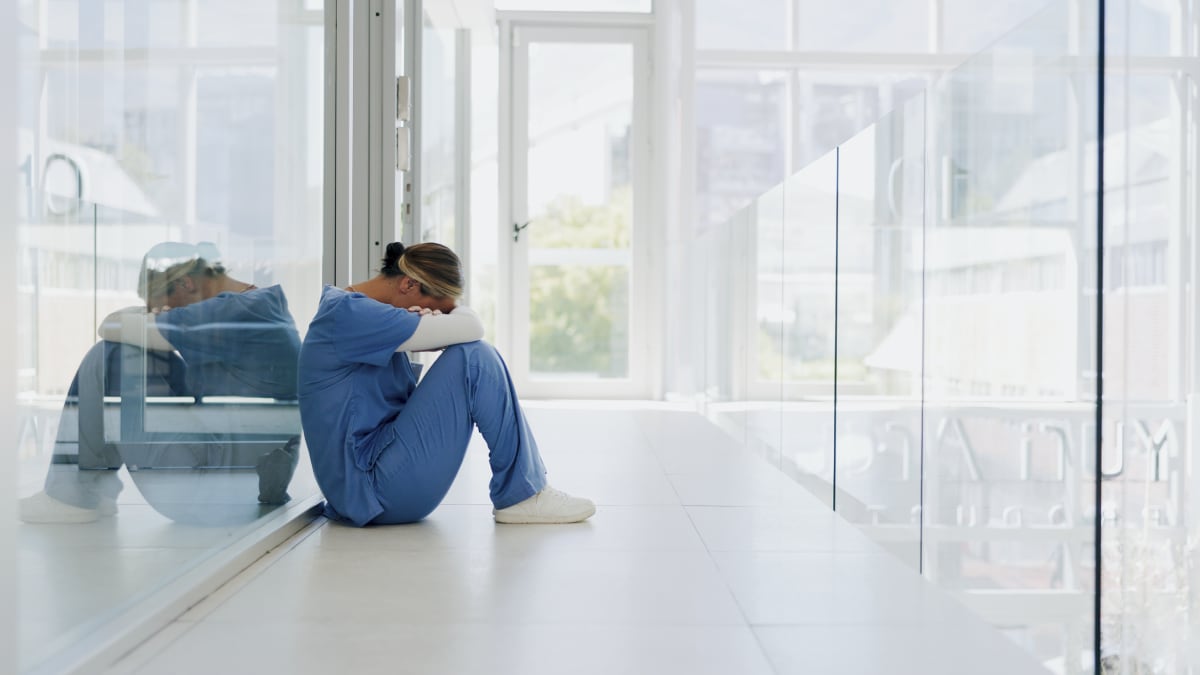 The image shows a clinician sitting in a hallway resting their head on their forearms.