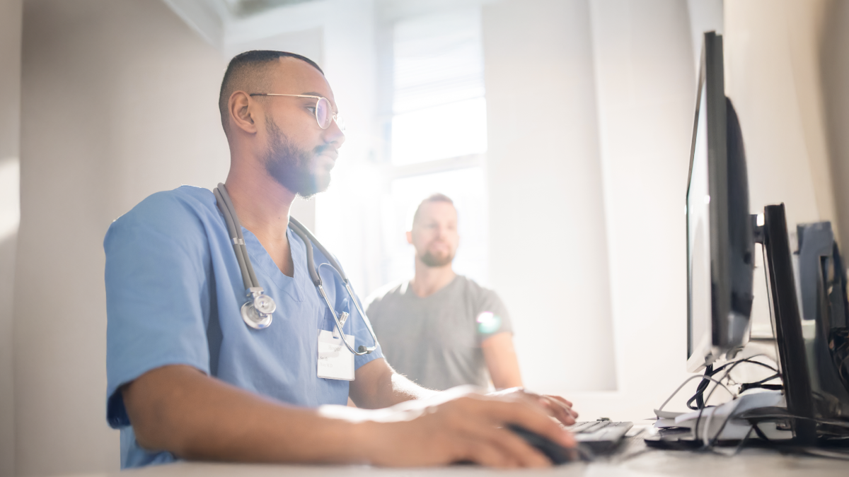 The image shows a clinician on a computer with a patient in the background.