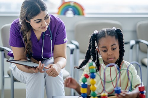 A young girl sits in a waiting room playing with a bead maze as her healthcare provider observes her playing and talks with her. The healthcare provider is dressed professionally in scrubs and has a tablet in hand as she takes notes.