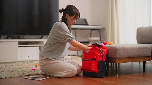 Woman kneels on floor while preparing red emergency bag in her living room at home.