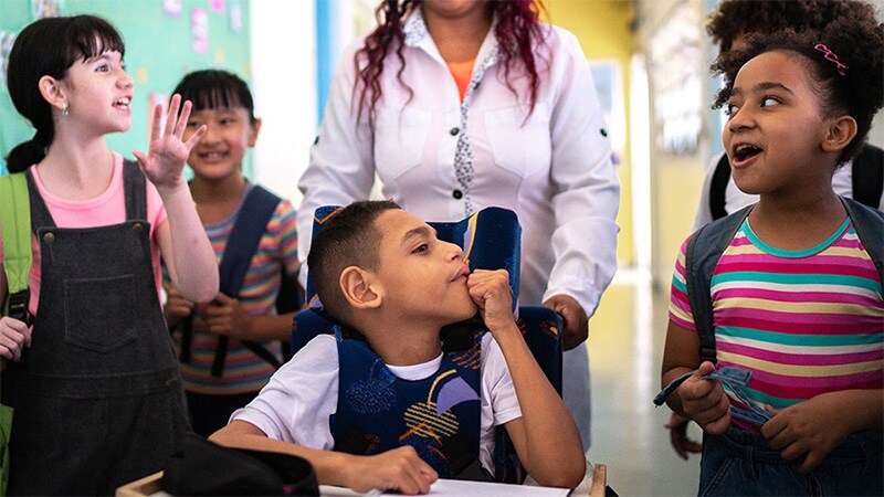 Assisted by a teacher, a child with cerebral palsy uses a wheelchair while talking with his friends in a classroom hallway.