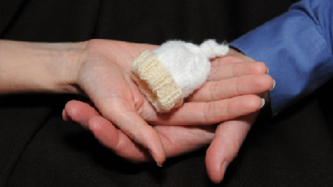 Two hands holding a tiny baby hat. Get 2023 stats on fetal mortality.