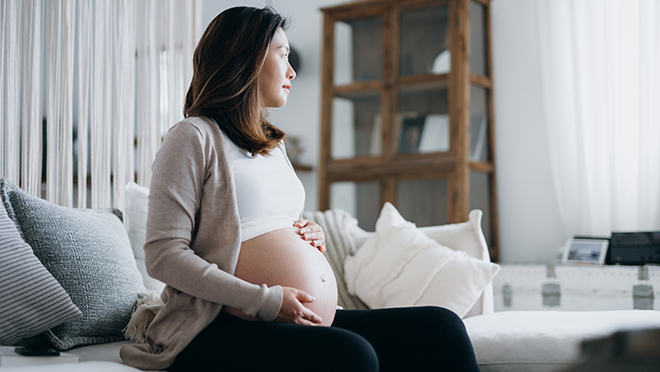 A pregnant woman sitting on a couch and rubbing her pregnant belly as she looks out the window.