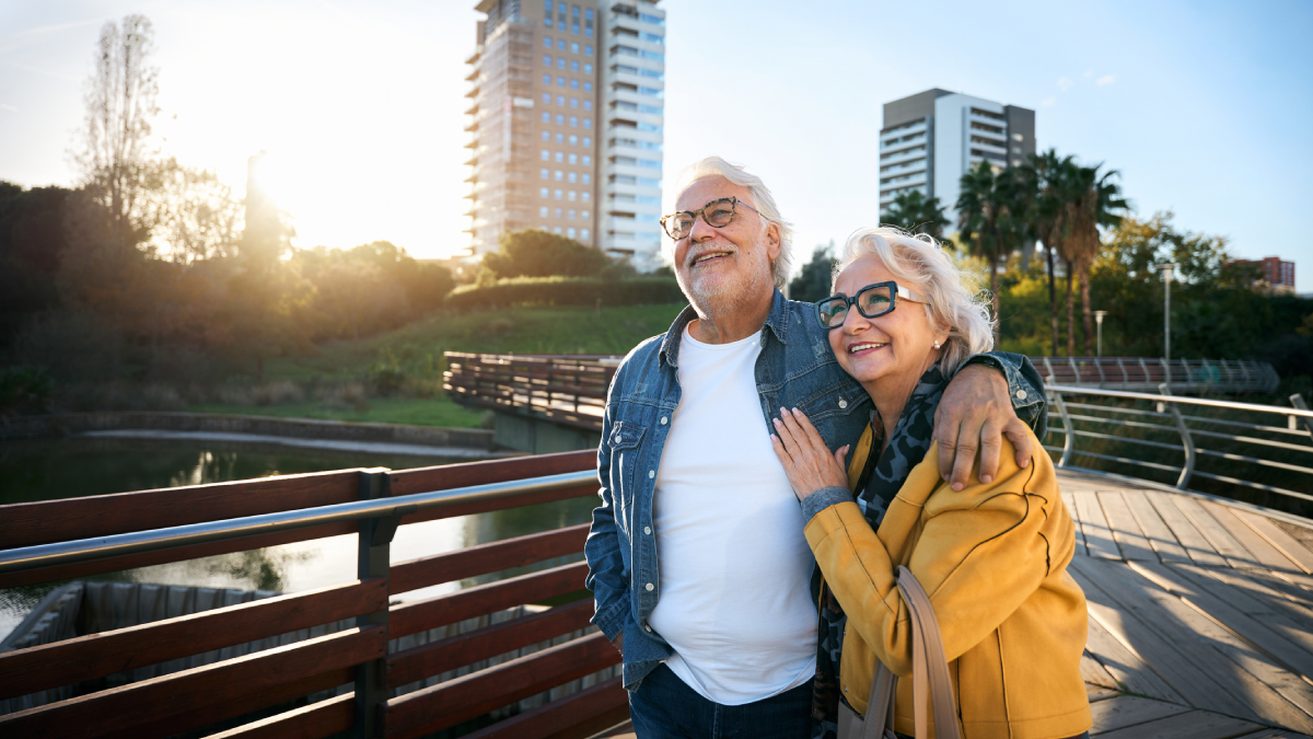 An older woman and man standing outside while embracing each other and looking off in the distance together, smiling.