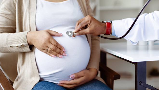 A woman holding her pregnant belly while a doctor listens with a stethoscope.