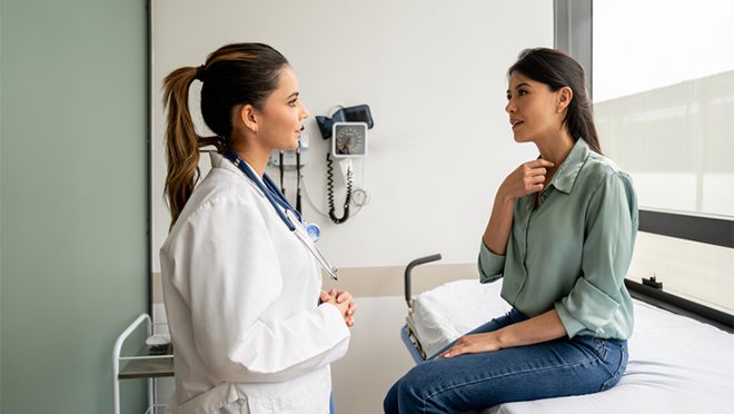 A woman sitting on a medical exam table talking to her doctor and gesturing to her throat.