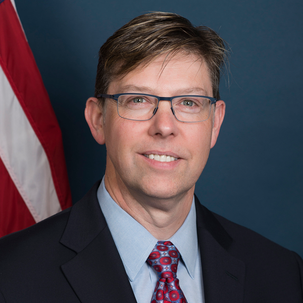 Middle-aged white man with blond hair and glasses. He wears a suit and tie in front of a blue background and U.S. flag.