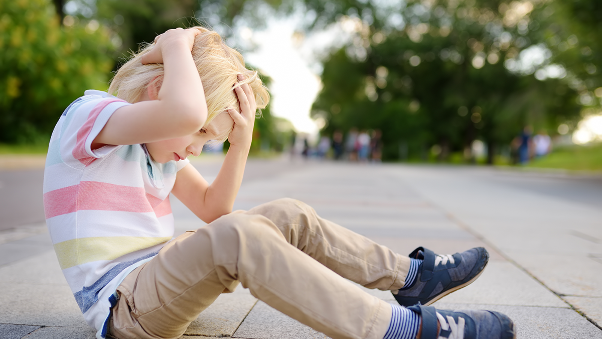 Young blond boy sits in tree-lined road clutching his head as if injured there