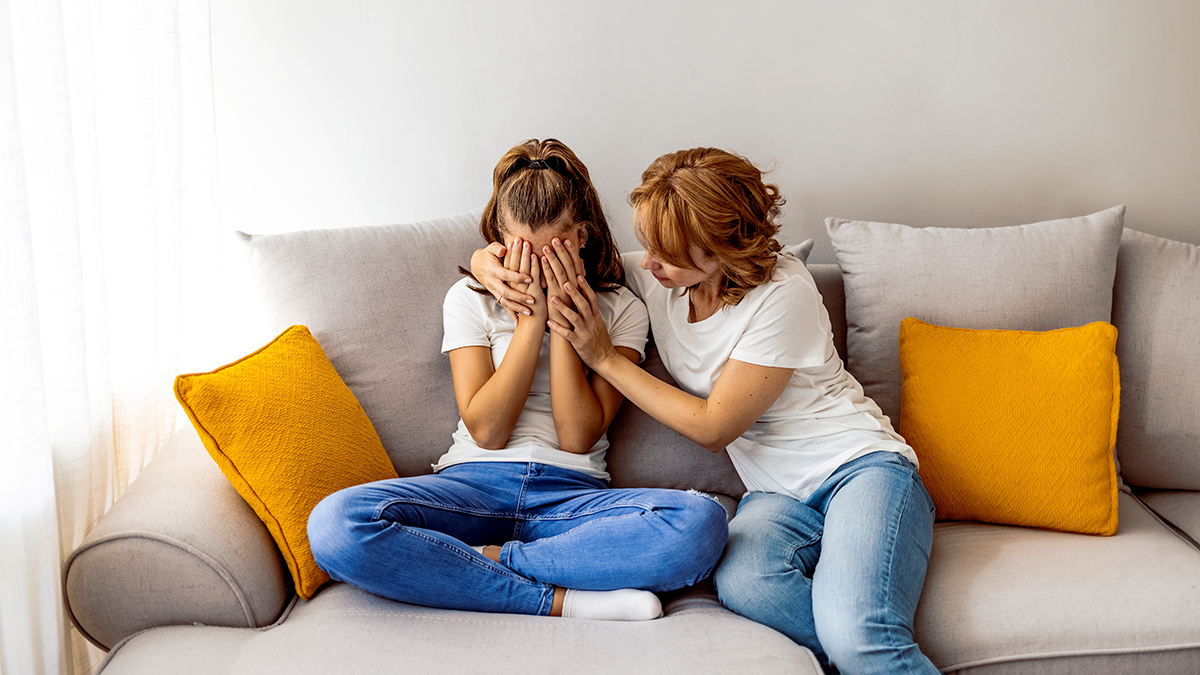 Teen and mom sit on couch. Teen cries while mom comforts her.