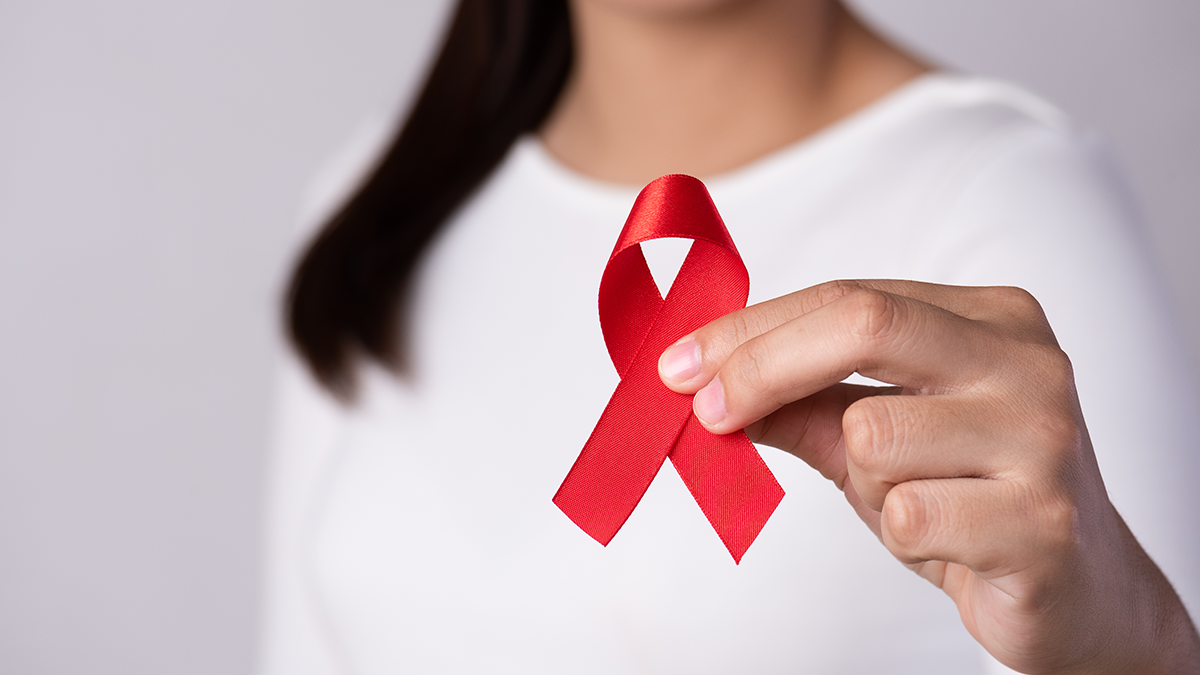 Torso, arms, and long dark hair of woman in white t-shirt holding red ribbon for HIV to camera
