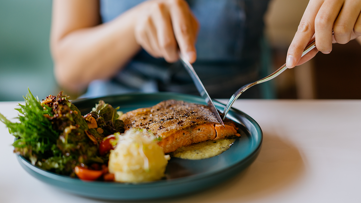 Person seated at table, image shows torso and hands as they eat meal of salmon, salad, and more