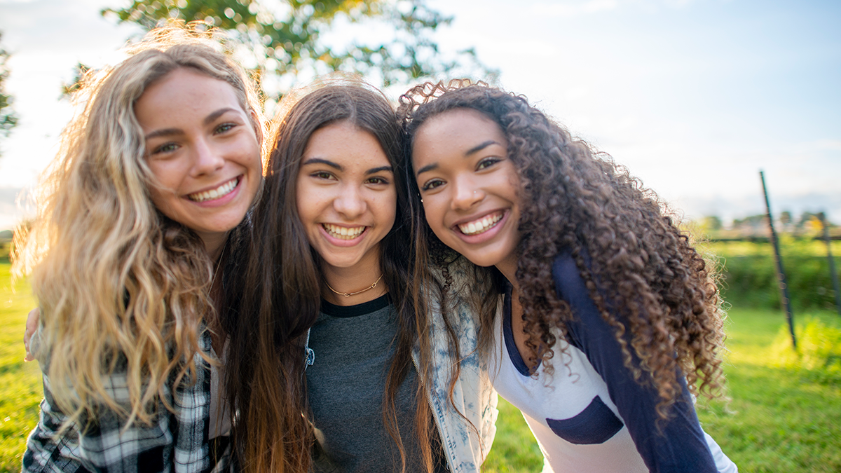 Three diverse teen girls lean into camera and smile in front of a field