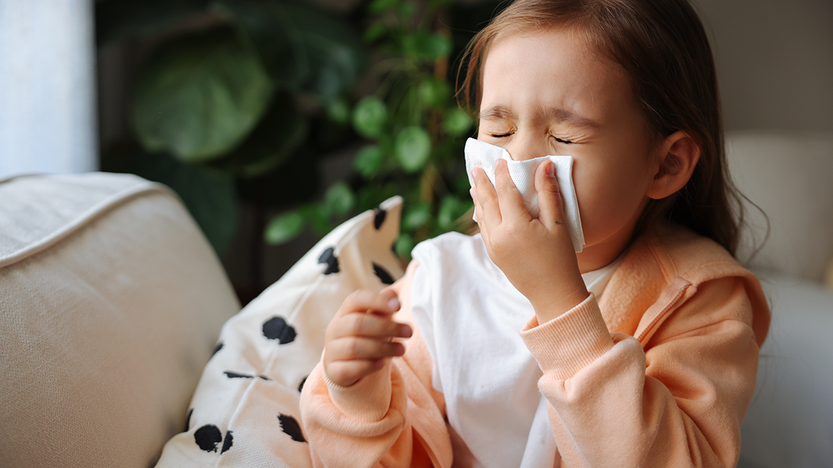 Little girl sits on couch and holds a tissue to her nose as she sneezes