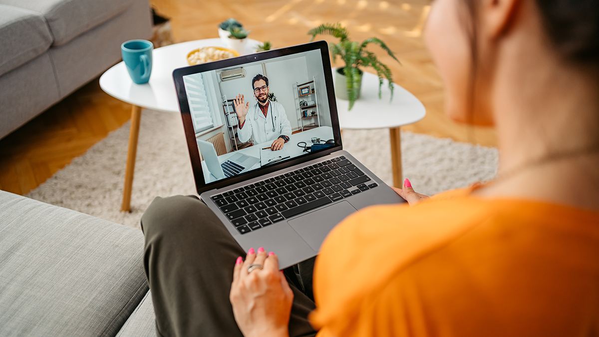 Woman sits with laptop on lap. On the screen is a doctor with whom she is consulting.