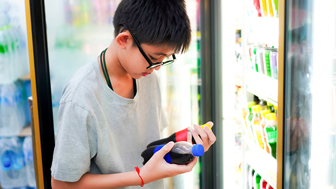 A young boy standing in front of a store refrigerator looking down at two sodas in his hands.