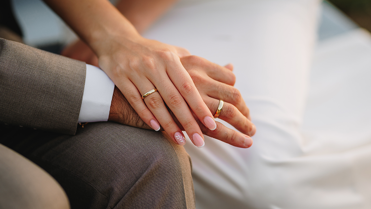 Close up of two hands. Man in suit; woman in wedding dress. Hands prominently display rings