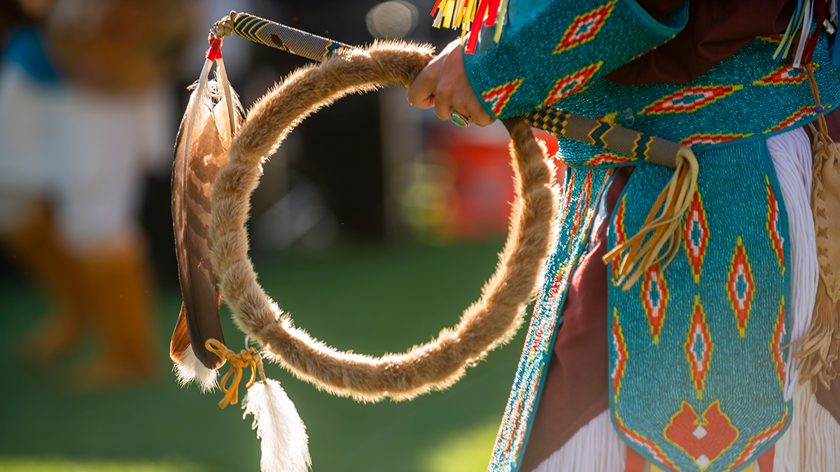 Person stands in native American clothing holding woven circle and stick with feathers