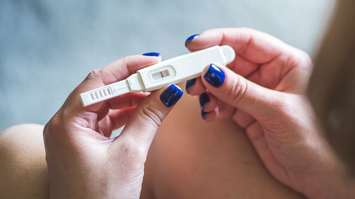 Hands with blue polish belonging to a young girl rest on knees and hold a positive pregnancy test