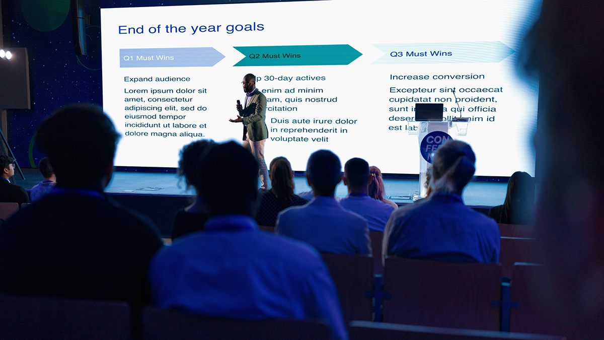 Group of conference attendees sit in the dark watching a presentation about end-of-year goals