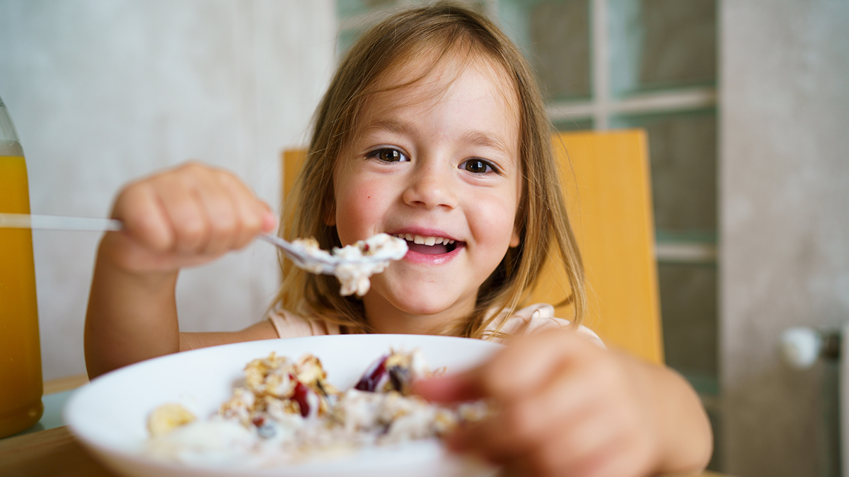 Smiling female child sits at table eating yogurt and granola from a bowl