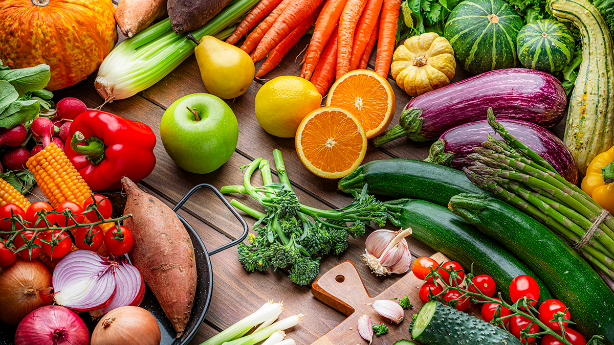 Table is covered with fruits and vegetables, including apples, oranges, peppers, eggplant, squash