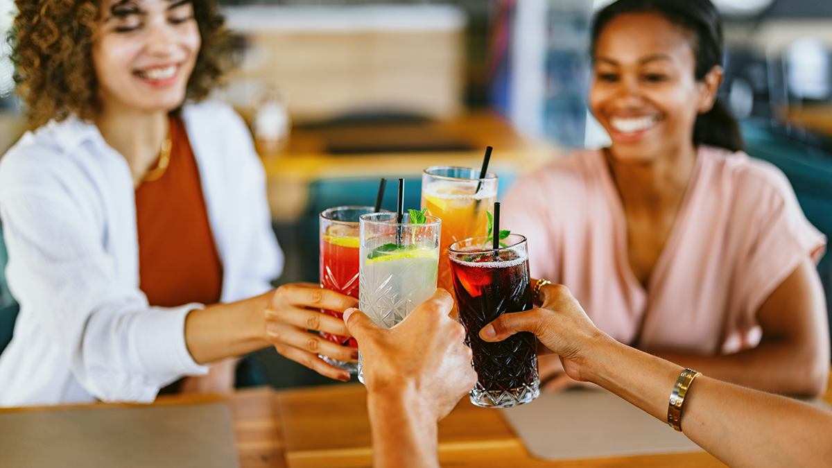 Four arms (two from visible women) toast with soda, tea, and lemonade