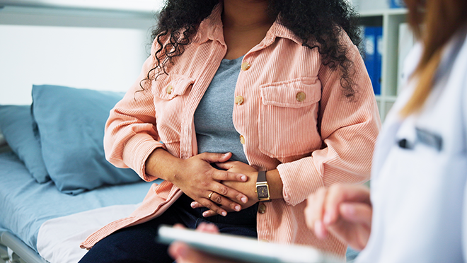 A cropped photo below the neck of a woman seeing her doctor and holding her lower abdomen.