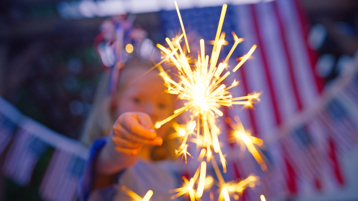 Young girl stands in front of a string of small American flags and one large one, holding a sparkler