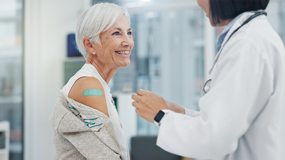 Older woman with white hair showing a teal bandage on her arm and smiling at a doctor wearing in white coat