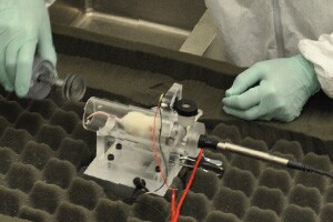 A white mouse rests in a small clear tube that is attached to a piece of metal (a plethysmograph). The tube is open at one end and the researcher%26rsquo;s gloved hand moves toward the tube with a silver tube-shaped stopper. The other end of the tube is sealed, with a plastic and metal wire of about %26amp;frac12; inch in diameter attached to it. There are also some thin wire leads attached to the plethysmograph . The researcher is wearing a white disposable lab coat and blue single-use gloves. The plethysmograph sits on a table covered with egg crate foam.