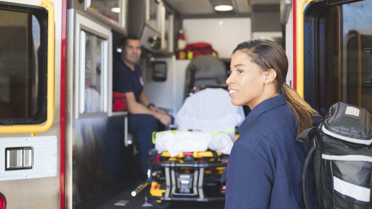 An EMT standing in the foreground in front of an empty stretcher inside of an ambulance.