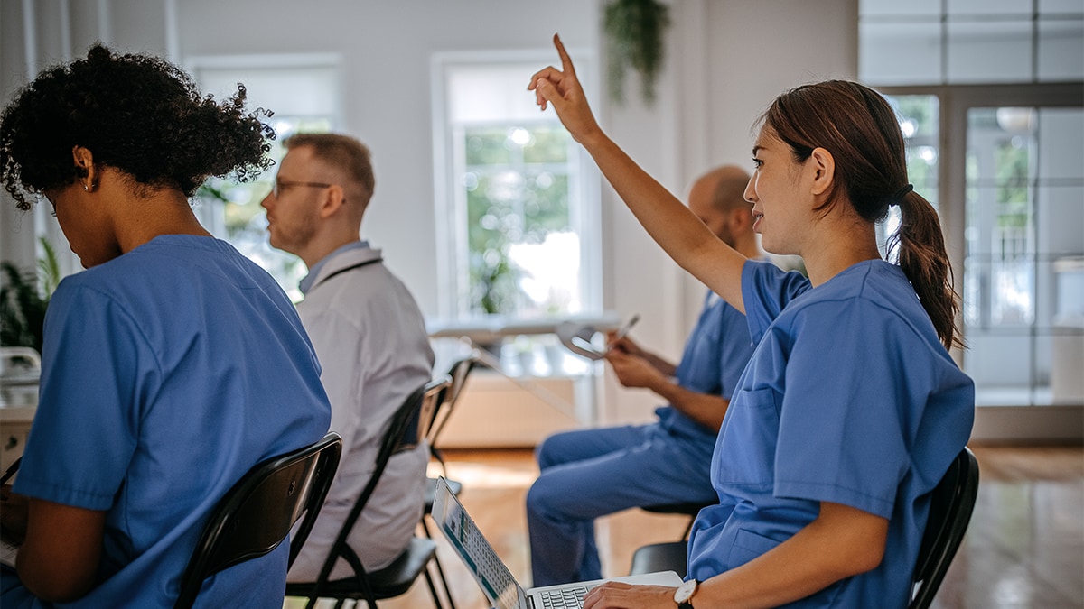 Woman in scrubs raises her hand at a healthcare worker town hall meeting.