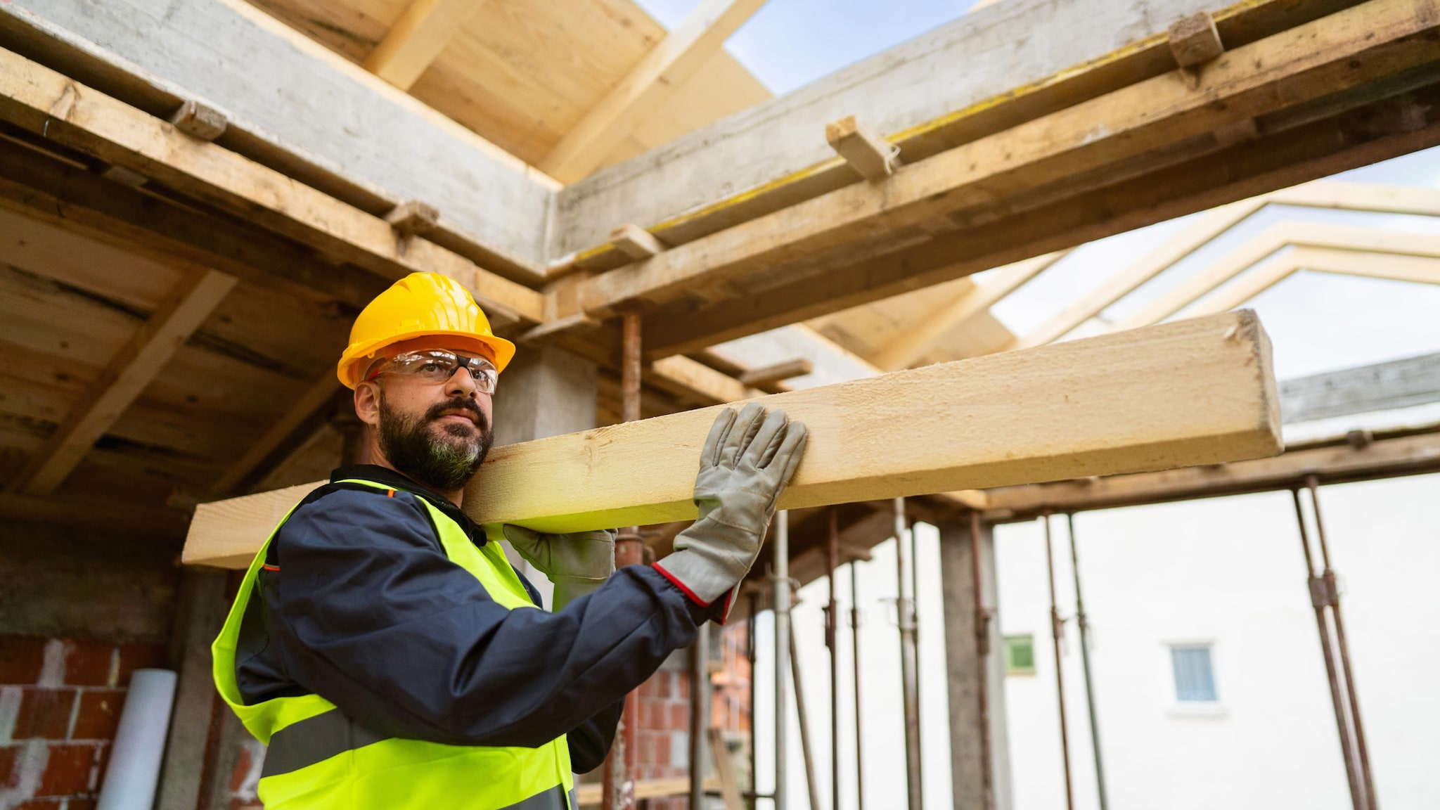 Man with a beard wearing a hard hat, carrying lumber at a construction site.