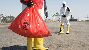 Waiste down picture of person wearing hazmat suit and carrying a bag marked as hazardous material.