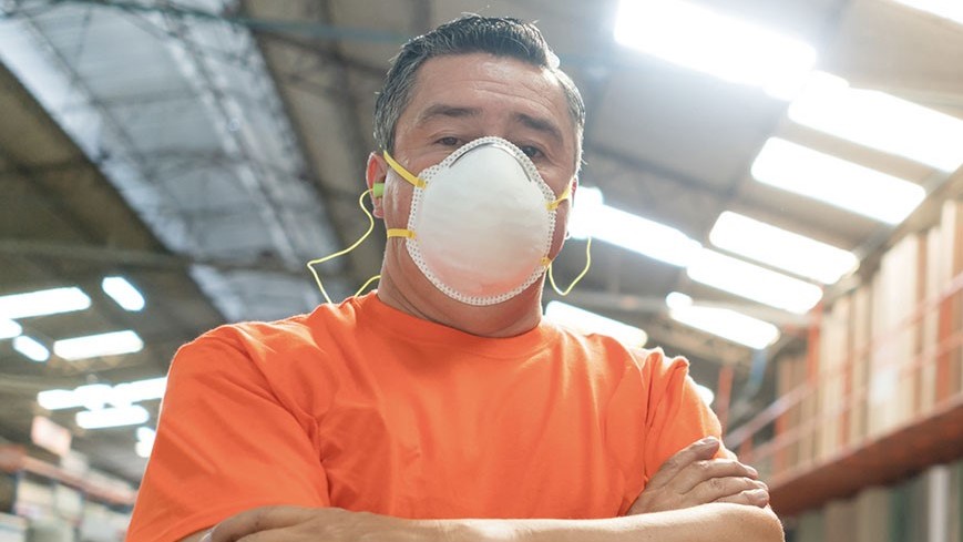 Worker looking at the camera with arms crossed wearing an FFR and hearing protection in a lumberyard.