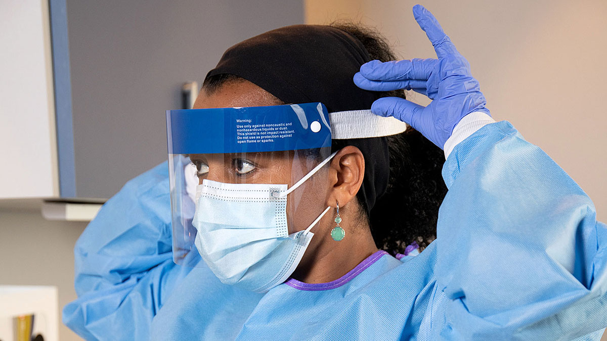 An African American Woman working in a medical setting wearing a surgical mask, gown, gloves and face shield.