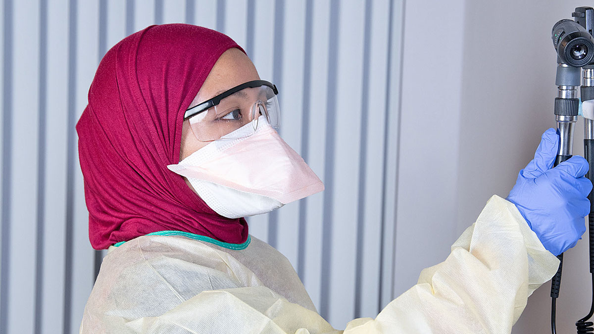 Asian woman wearing a filtering face respirator (duckbill), gown, head scarf (hijab), gloves, and goggles.