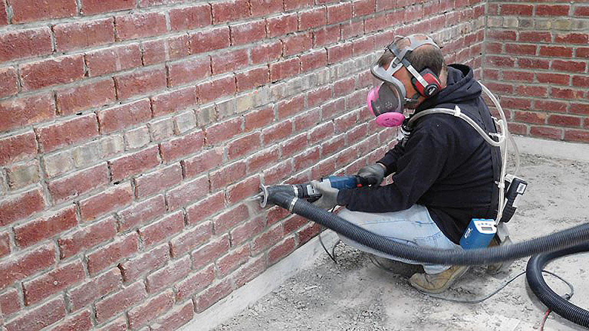 A worker kneeling down while tuckpointing with dust controls and respirator.