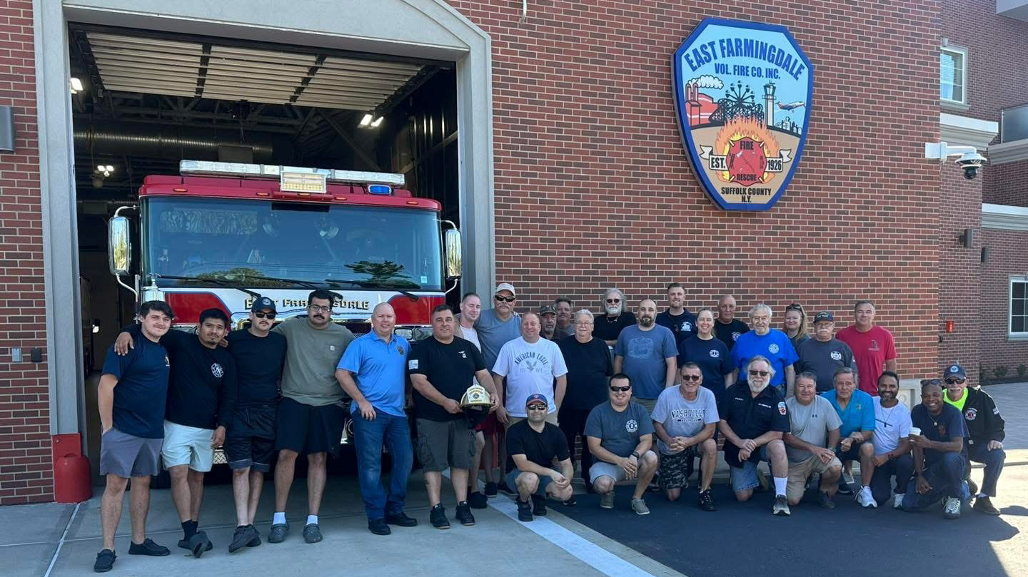 Group of firefighters standing in front of fire house and truck with one holding a gold helmet