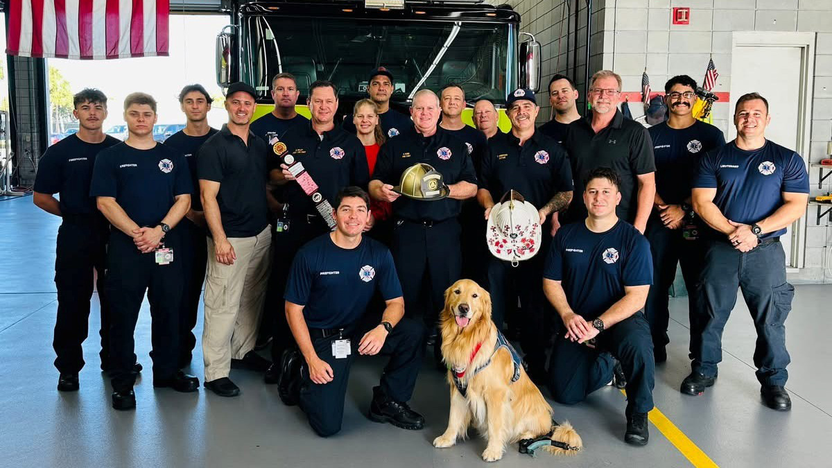 Group of firefighters inside firehouse in front of a truck posing with a gold helmet and golden retriever