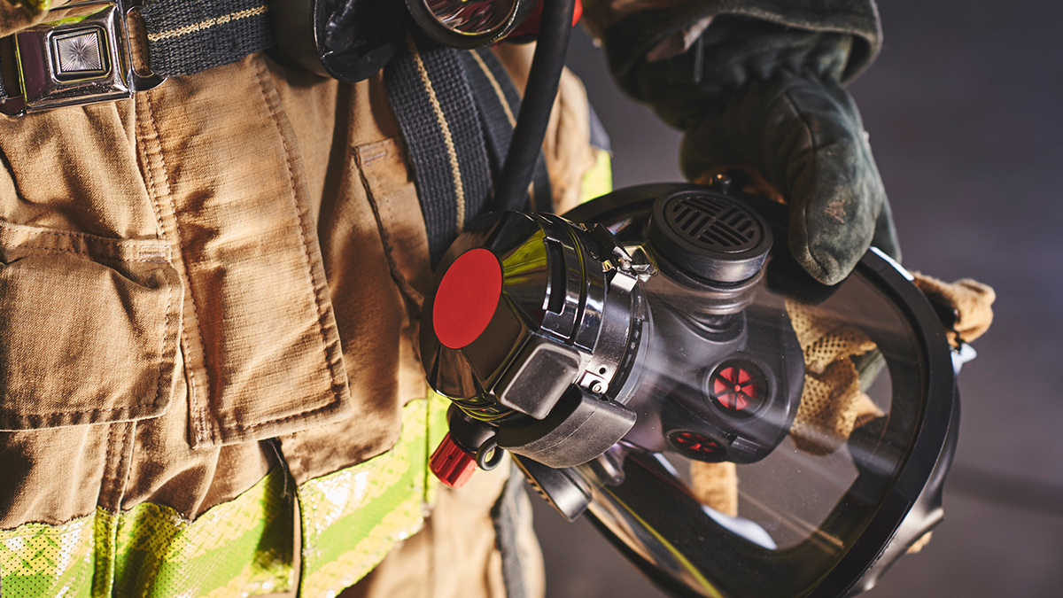 Close-up of a firefighter holding a protective breathing mask, wearing tan turnout gear with reflective safety stripes and heavy gloves.