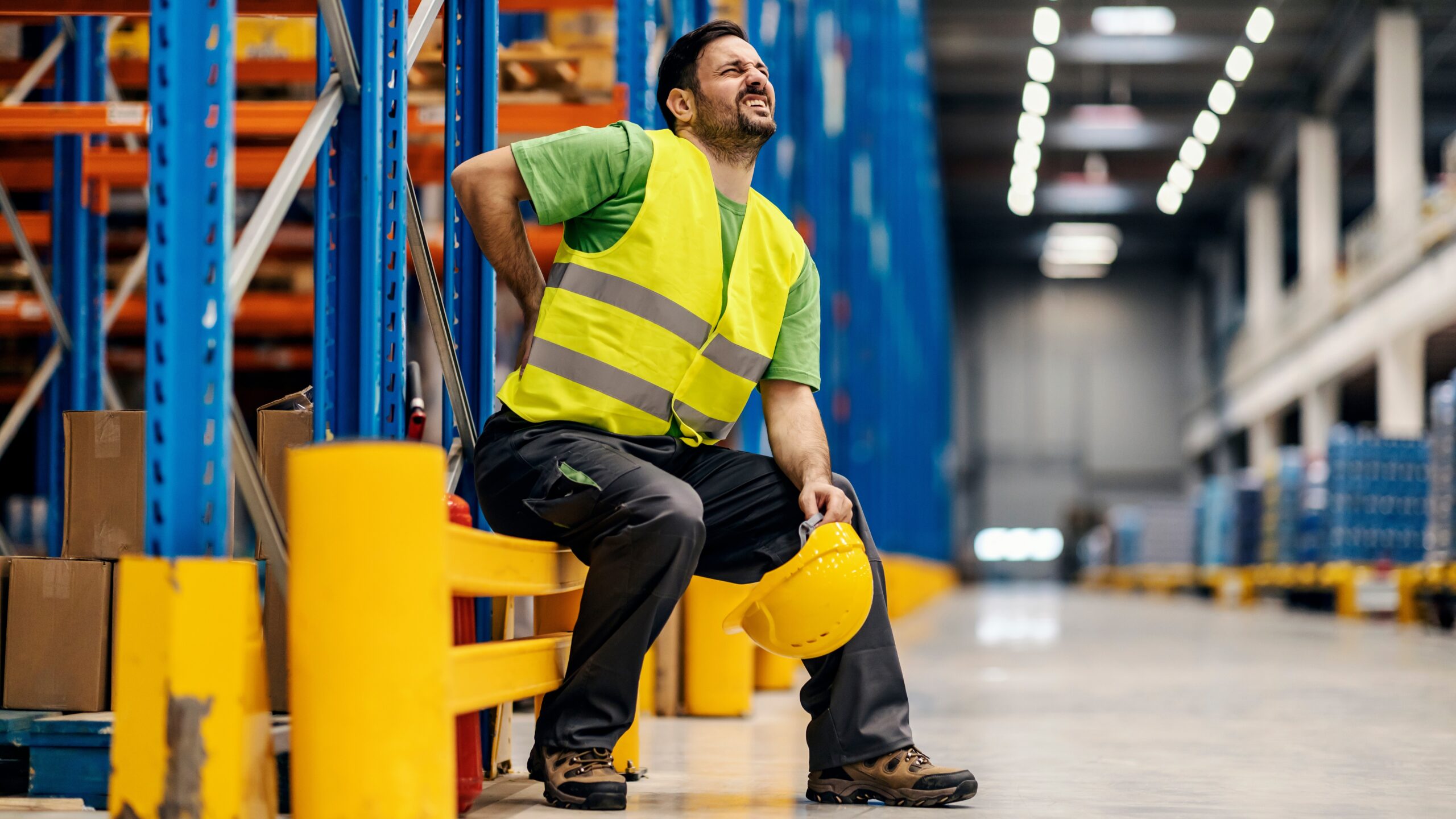 Man in safety vest sitting down, rubbing his back with a pained face